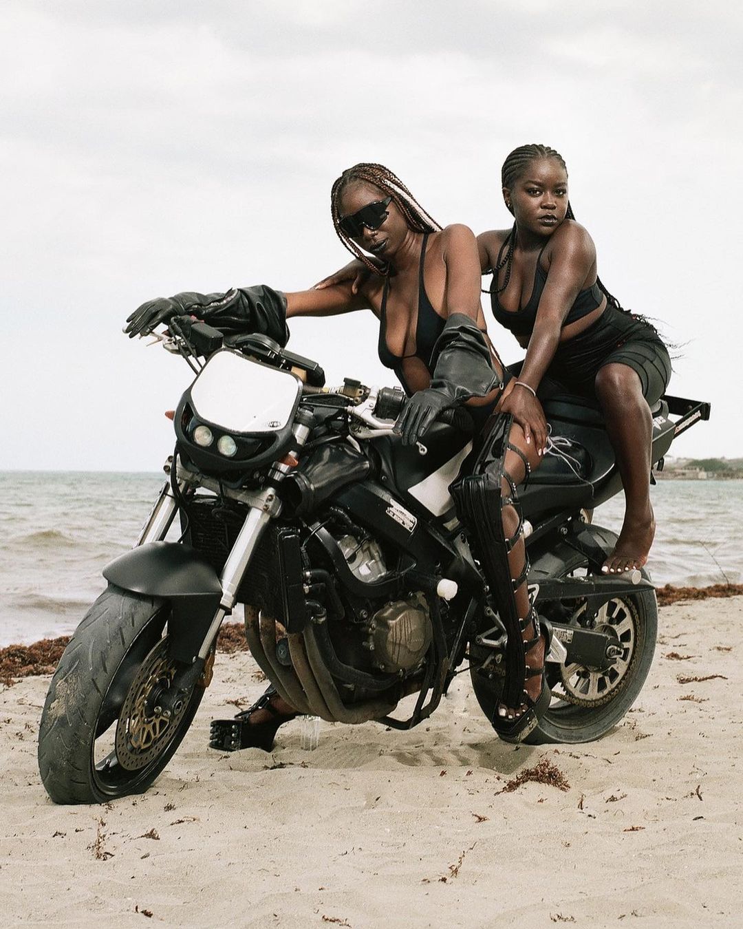 Two women pose on a black motorcycle on a beach with sunglasses and black clothing, sea and sky visible in the background.