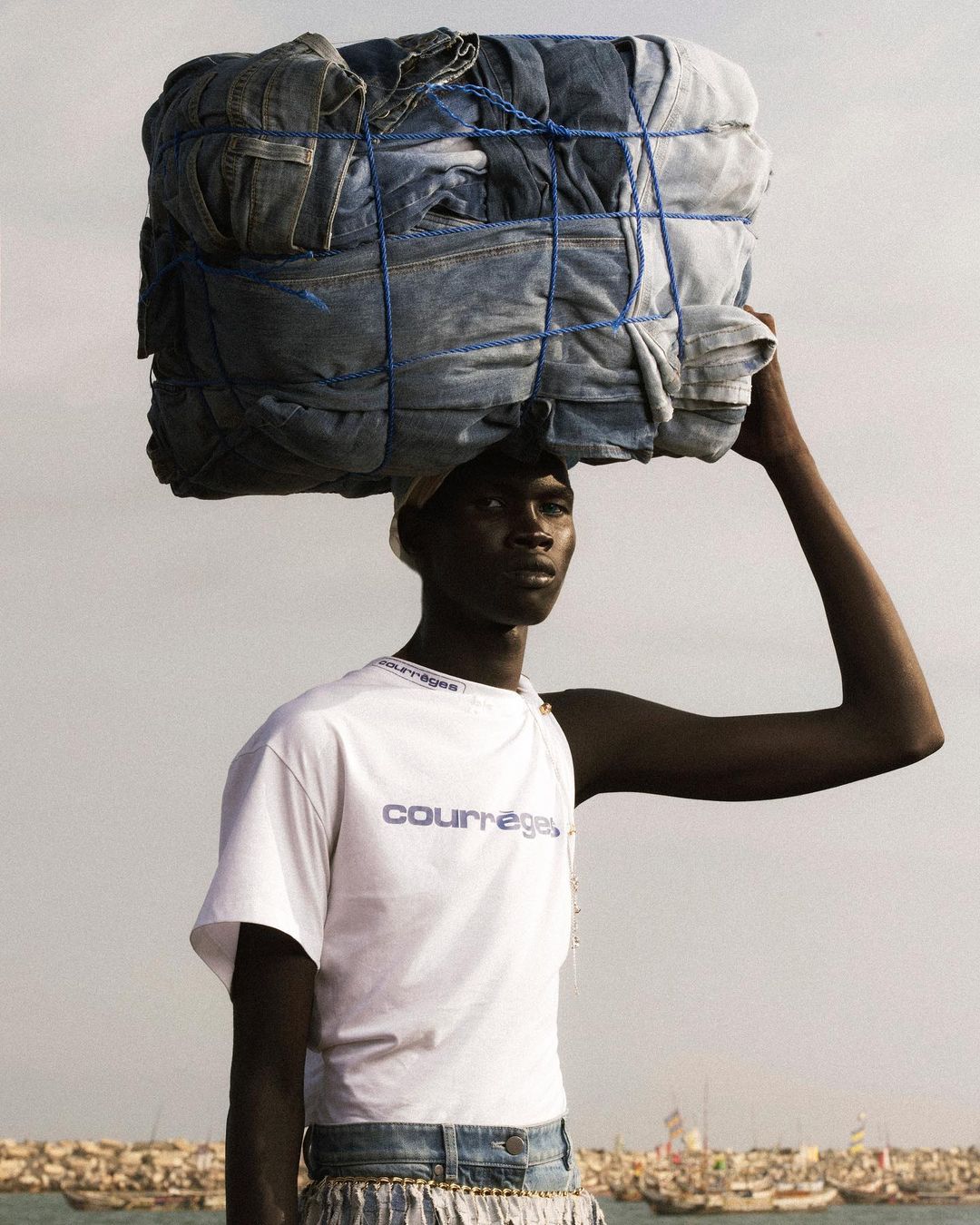 Dark-skinned adult male carries a stack of blue jeans on his head near the ocean in an outdoor fashion editorial shot.