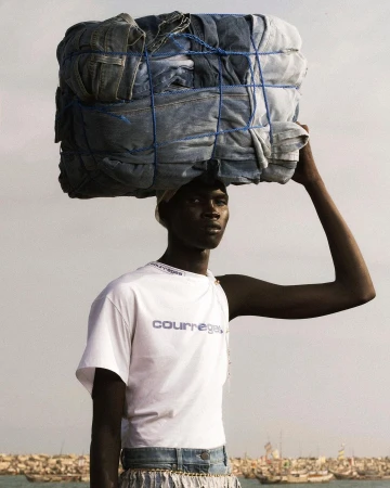 Dark-skinned adult male carries a stack of blue jeans on his head near the ocean in an outdoor fashion editorial shot.