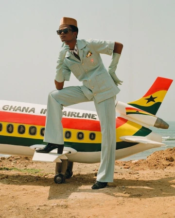 A dark-skinned adult male in a pale-blue suit and gloves poses on a Ghana International Airlines airplane prop outdoors on a sunny day.