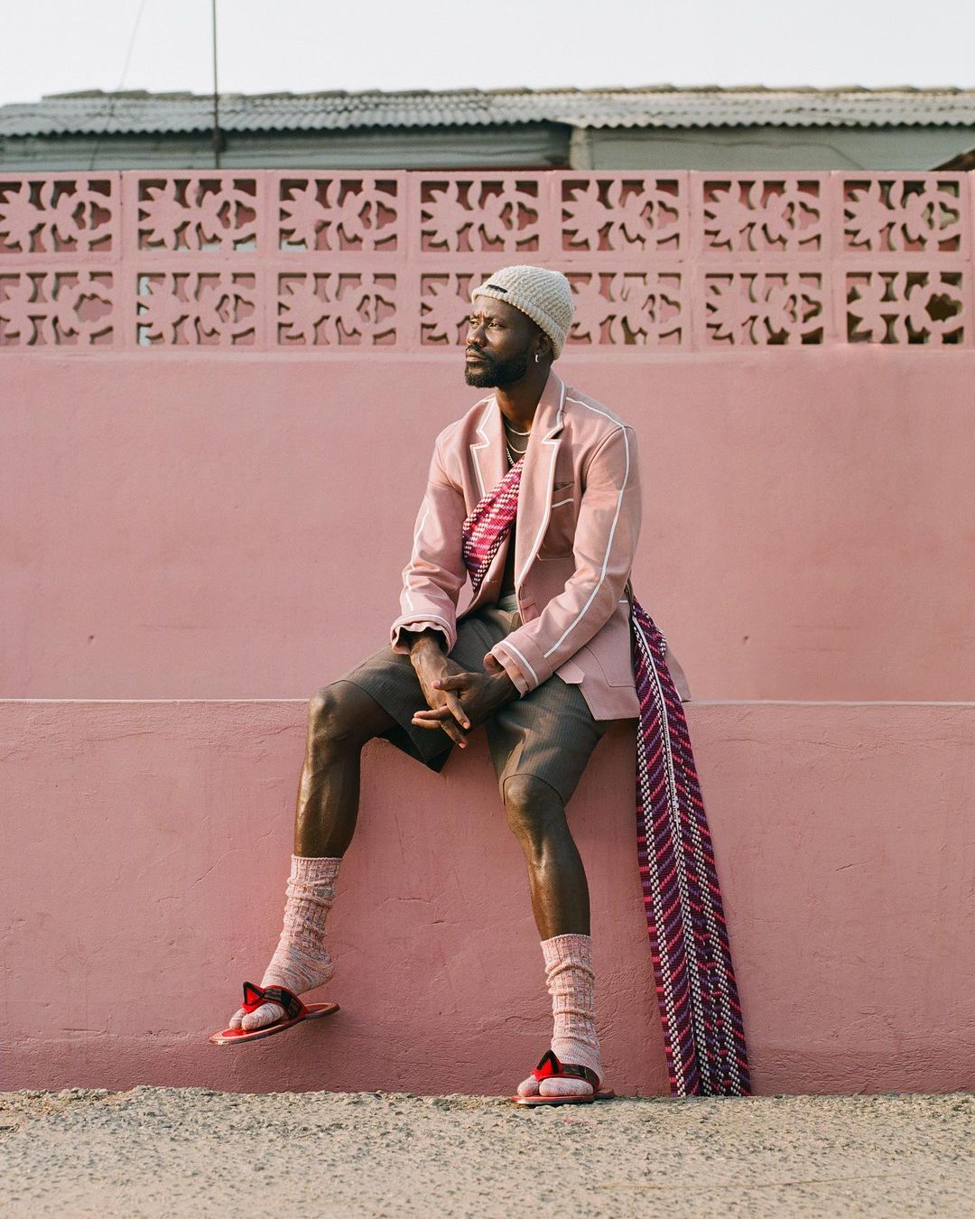 Man in pink blazer and shorts sits on a pink wall wearing a cream beanie, socks, and sandals in an outdoor daytime setting.
