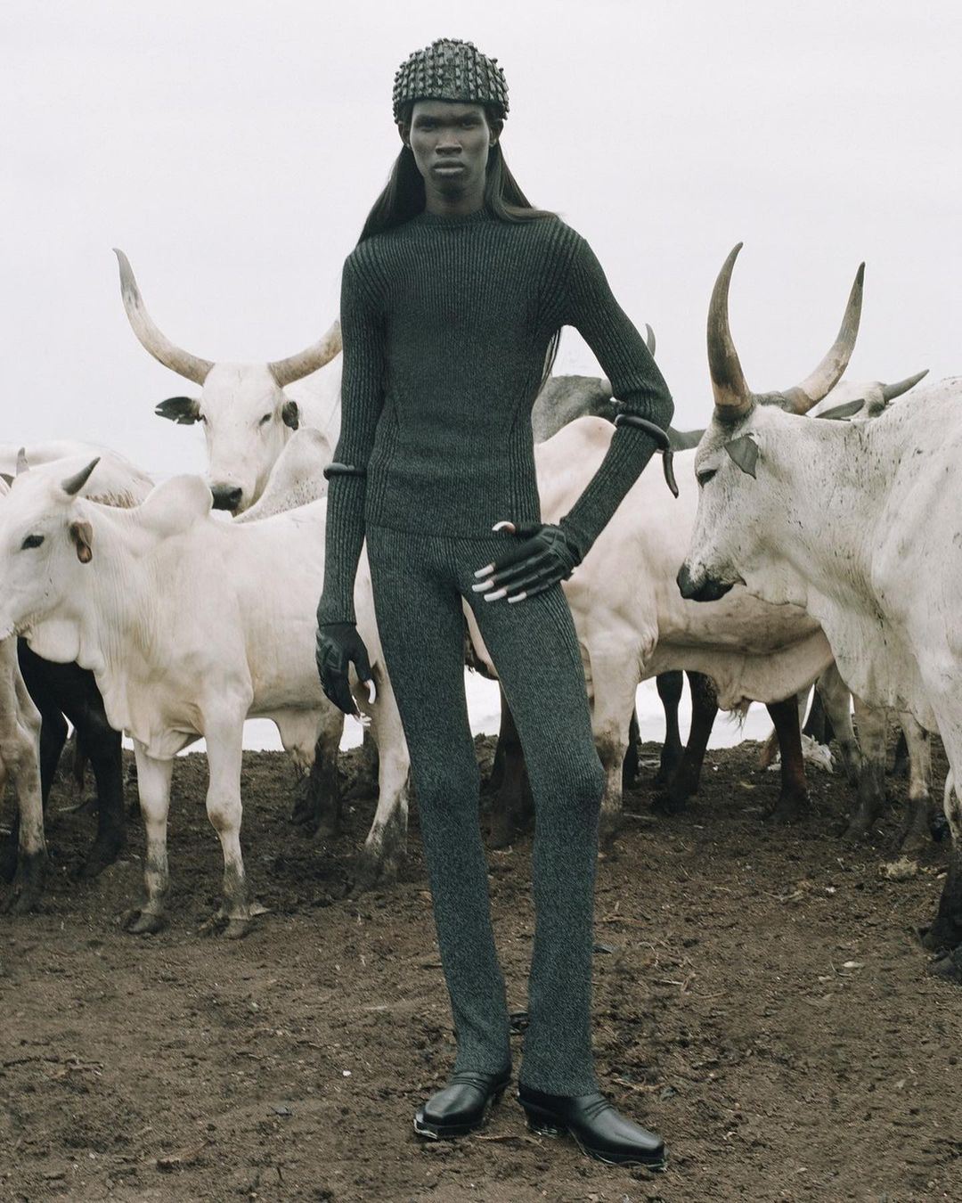 Dark-skinned person in a knit suit poses in a field with white, horned cattle against a pale sky in a full shot, fashion-editorial style.
