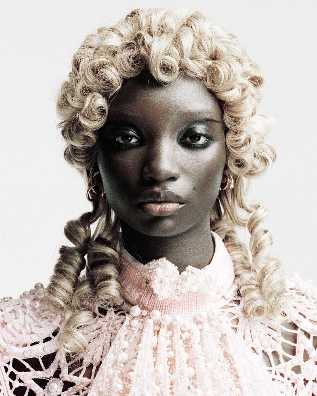 Close-up studio portrait of a Black woman with ornate blonde wig and pastel pink, lacy, beaded dress against a neutral backdrop.
