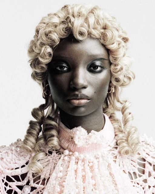 Close-up studio portrait of a Black woman with ornate blonde wig and pastel pink, lacy, beaded dress against a neutral backdrop.
