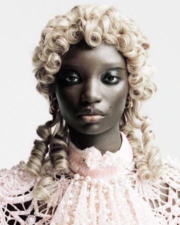 Close-up studio portrait of a Black woman with ornate blonde wig and pastel pink, lacy, beaded dress against a neutral backdrop.
