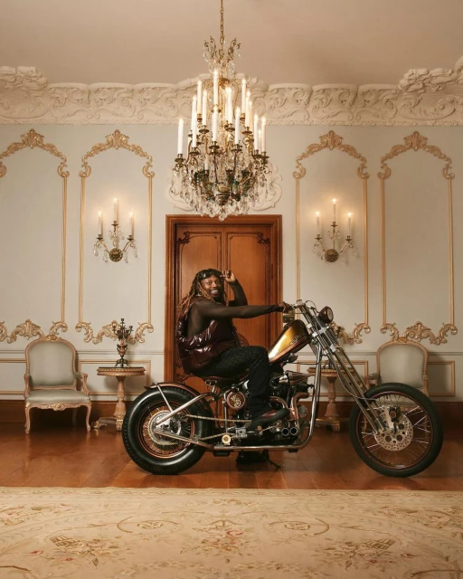 Stylish adult male with dreadlocks sits on a gold custom chopper motorcycle in an ornate, light-filled interior with decorative walls.