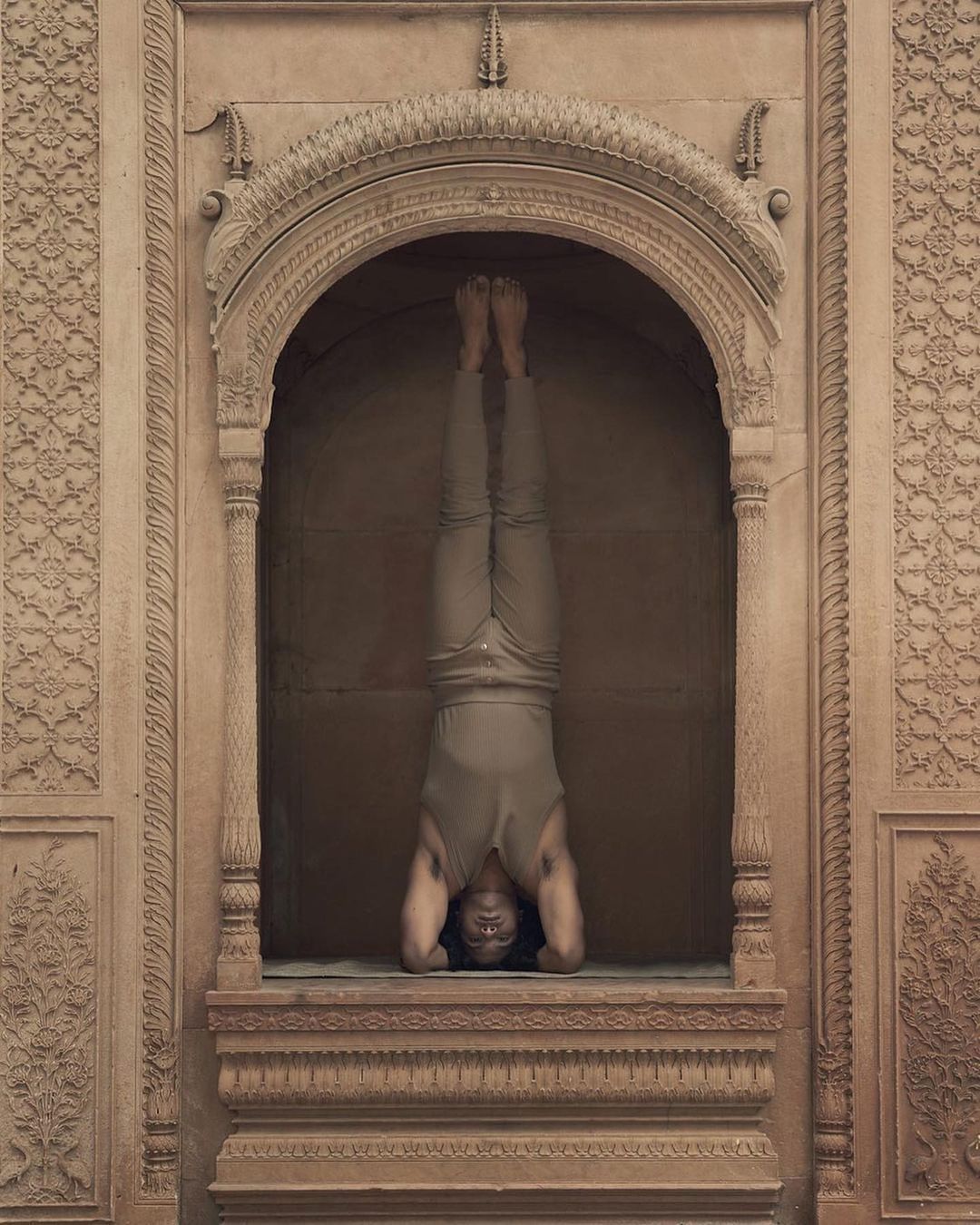 Adult male performing headstand yoga pose in stone archway. Man in a sleeveless shirt, pants and bare feet is balanced upside down.