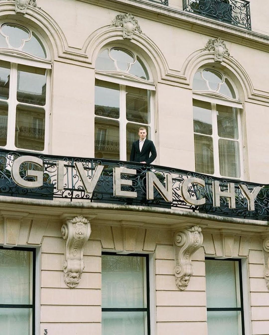 A man stands on a balcony with the GIVENCHY logo in Paris, wearing a suit in an outdoor setting with a building facade in the background.
