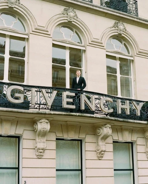 A man stands on a balcony with the GIVENCHY logo in Paris, wearing a suit in an outdoor setting with a building facade in the background.