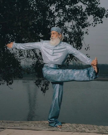 Elderly man with white beard wearing sparkly blue shirt and pants practicing yoga outdoors by a river, photographed by Kenny Germé