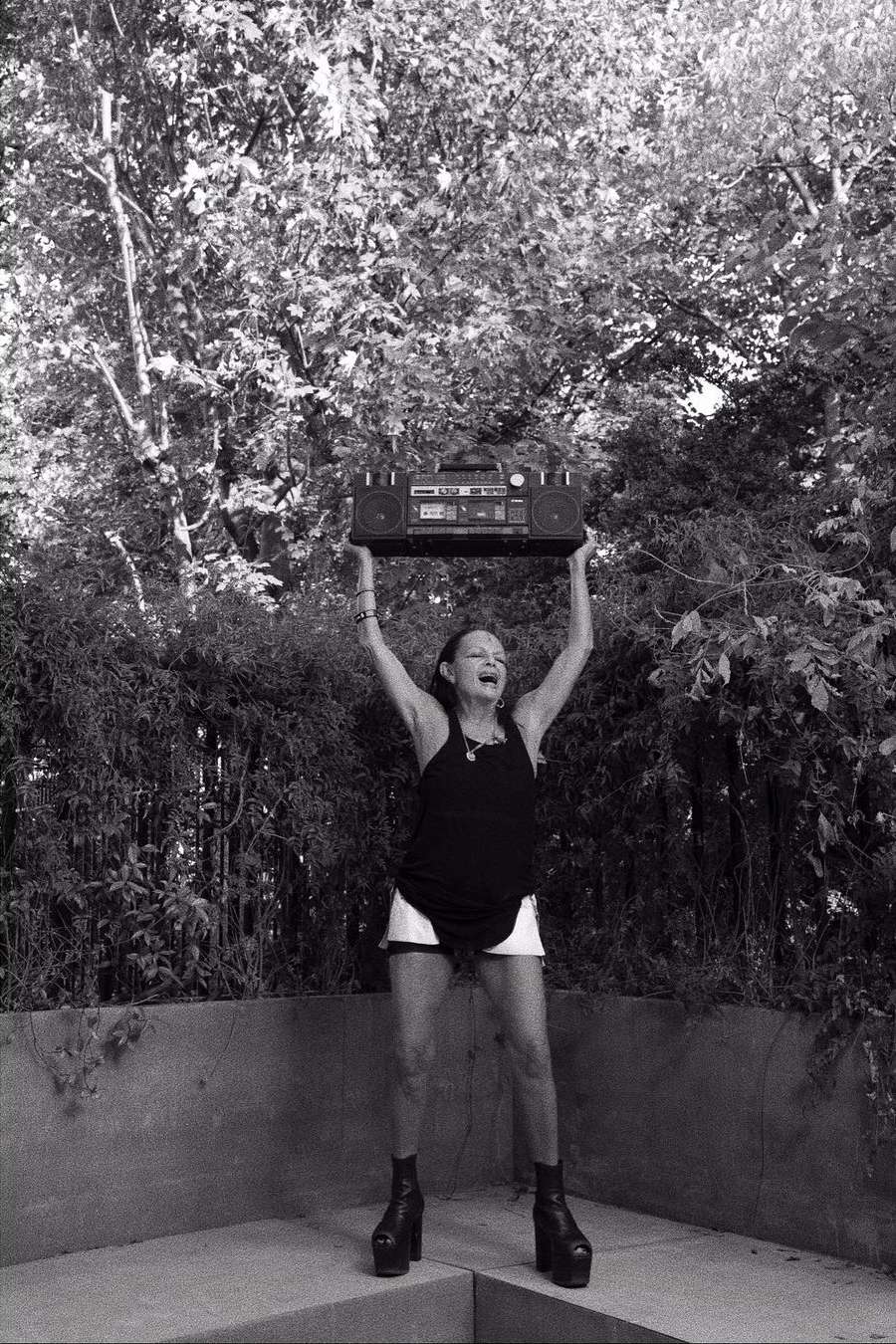 Monochrome shot of an adult woman holding a boombox overhead in an outdoor setting with lush greenery in the background. She's wearing a dark top and light shorts.