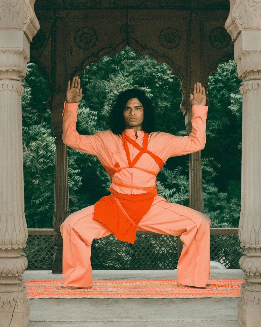 Male model in a peach-colored outfit strikes a pose in an arched, stone structure surrounded by green trees, showcasing fashion and style.