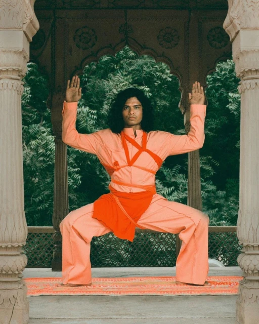 Male model in a peach-colored outfit strikes a pose in an arched, stone structure surrounded by green trees, showcasing fashion and style.