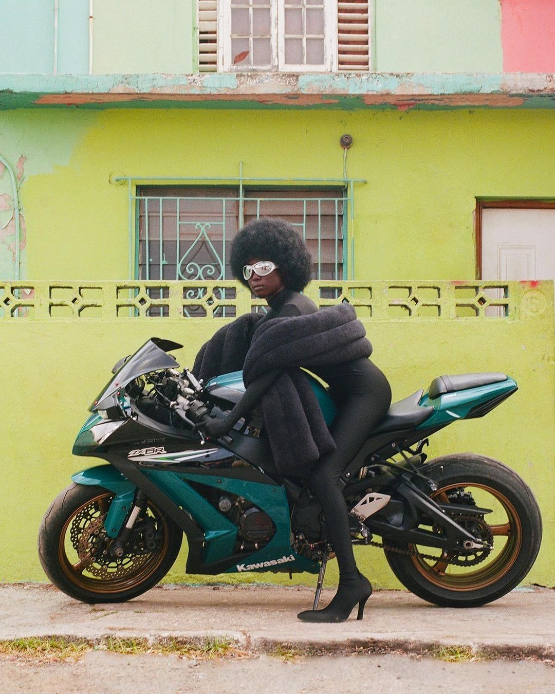 A woman with an afro wearing a black jumpsuit, sunglasses, and heels is posed on a green and black Kawasaki motorcycle outdoors.