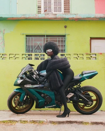 A woman with an afro wearing a black jumpsuit, sunglasses, and heels is posed on a green and black Kawasaki motorcycle outdoors.