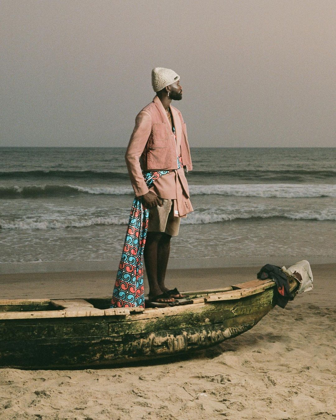 Man in pink suit stands in weathered boat on sandy beach near ocean under overcast sky, holding patterned textile; fashion editorial shot.