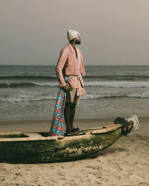 Man in pink suit stands in weathered boat on sandy beach near ocean under overcast sky, holding patterned textile; fashion editorial shot.