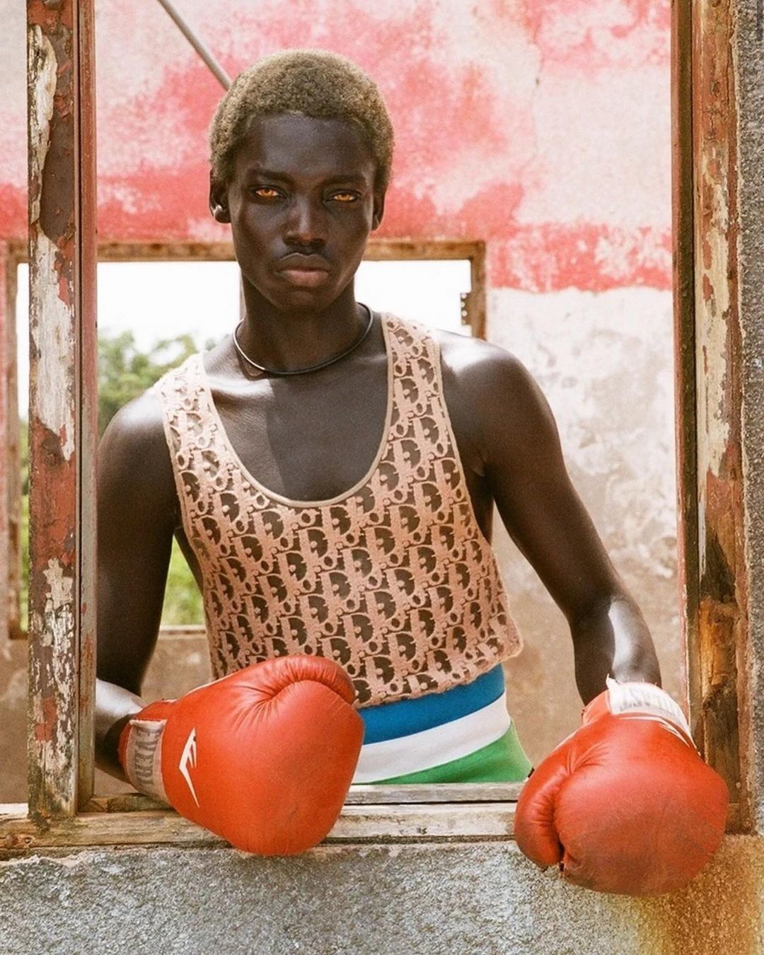 Portrait of a dark-skinned male athlete wearing a Dior tank top and red boxing gloves, standing in an abandoned building opening.