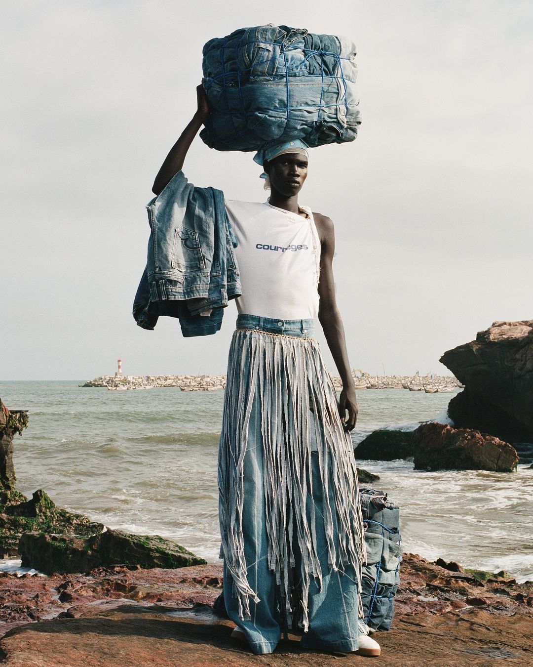 Fashion shot of a black model carrying a bale of denim on her head. She is wearing a white top and a long denim skirt with tassels.