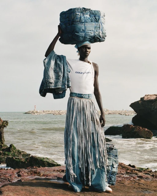 Fashion shot of a black model carrying a bale of denim on her head. She is wearing a white top and a long denim skirt with tassels.