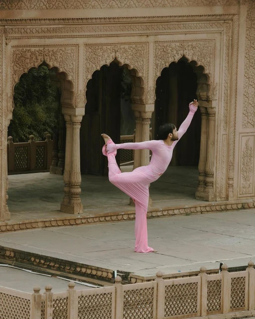 Adult in pink yoga suit balances in dancer pose on a stone terrace with arched openings in the background, serene outdoor setting.