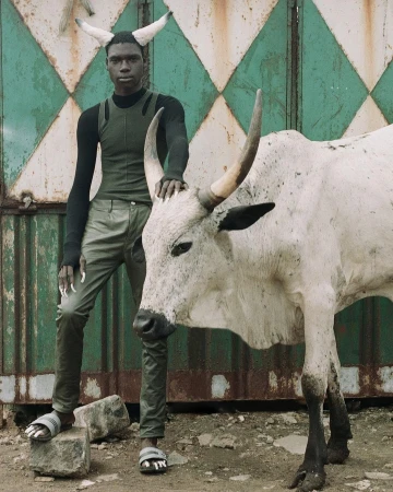 A person with horn-like prosthetics poses with a white cow in front of a green and white diamond-patterned metal wall outdoors.