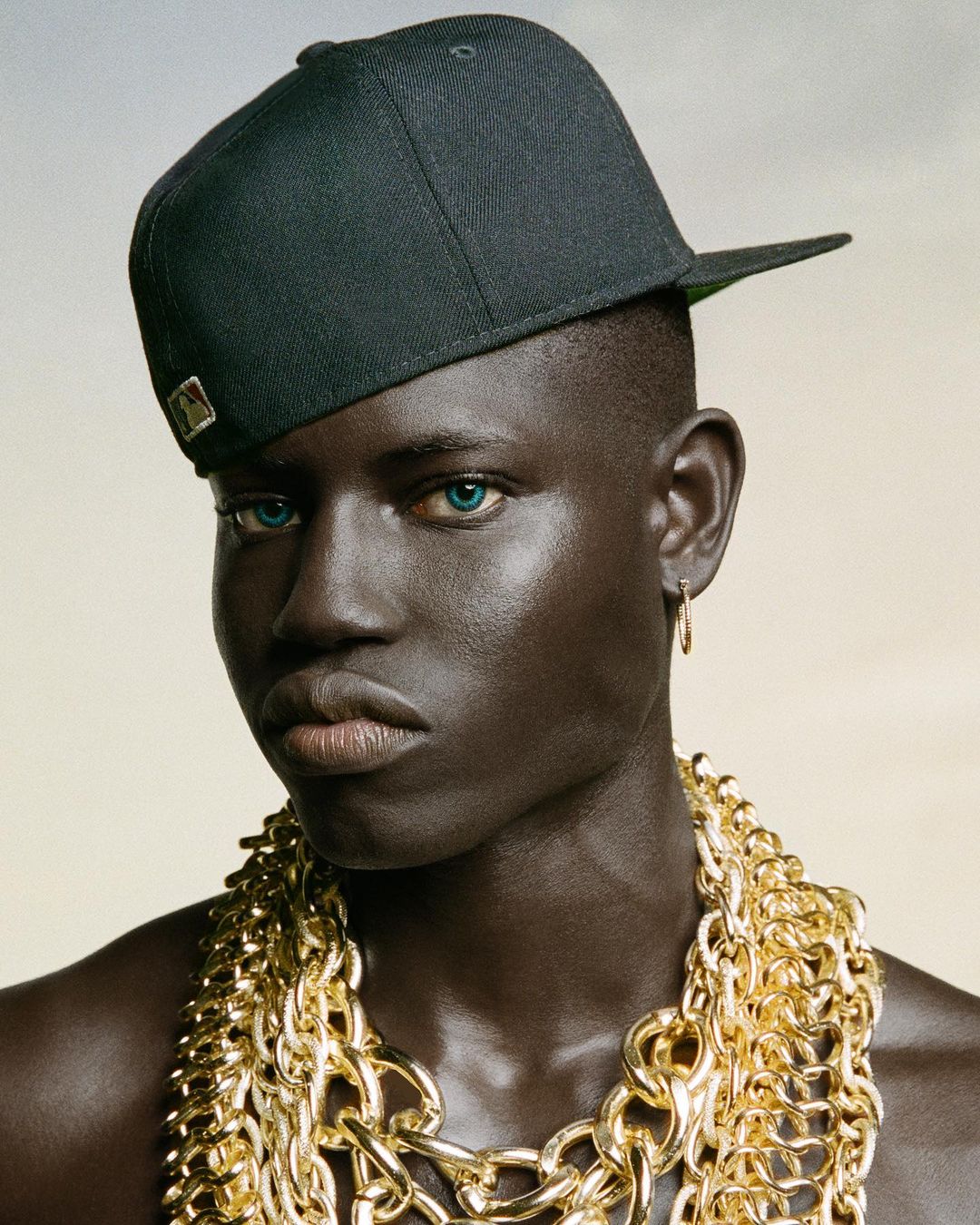 Headshot of a shirtless dark-skinned adult male with green eyes wearing a baseball cap and gold chain necklaces in a neutral studio.