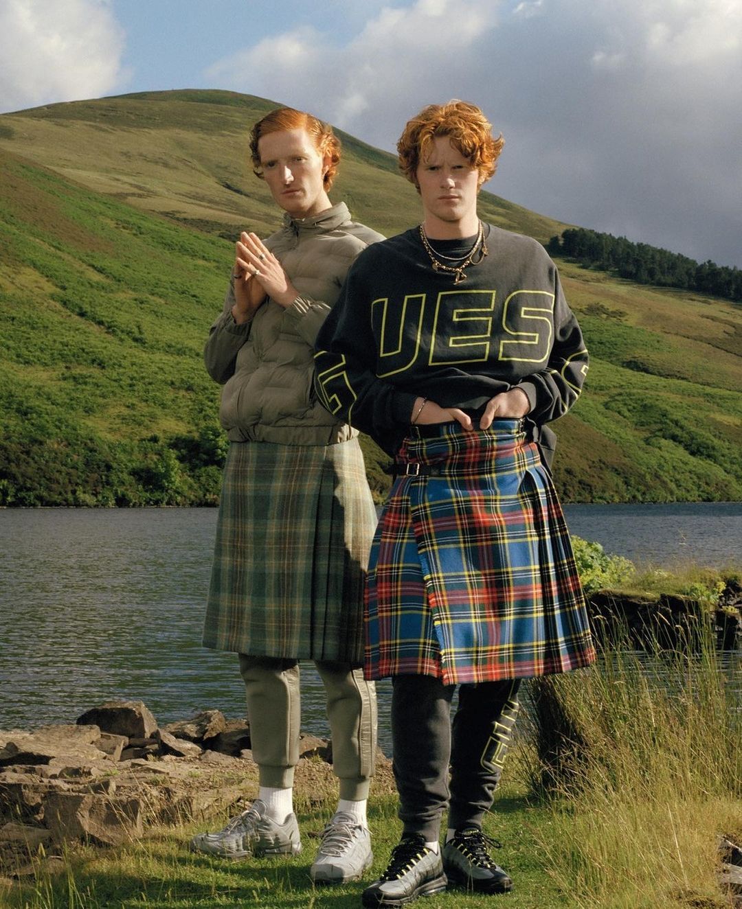 Two red-haired men wearing kilts stand in a grassy field by a loch, backed by rolling green hills under a blue sky with scattered clouds.