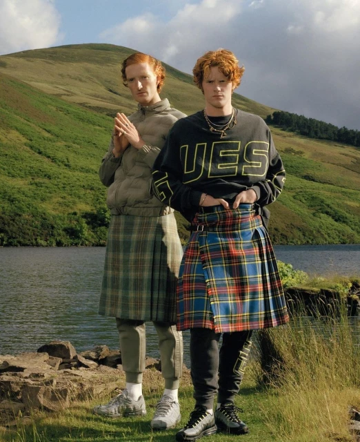 Two red-haired men wearing kilts stand in a grassy field by a loch, backed by rolling green hills under a blue sky with scattered clouds.