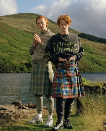 Two red-haired men wearing kilts stand in a grassy field by a loch, backed by rolling green hills under a blue sky with scattered clouds.