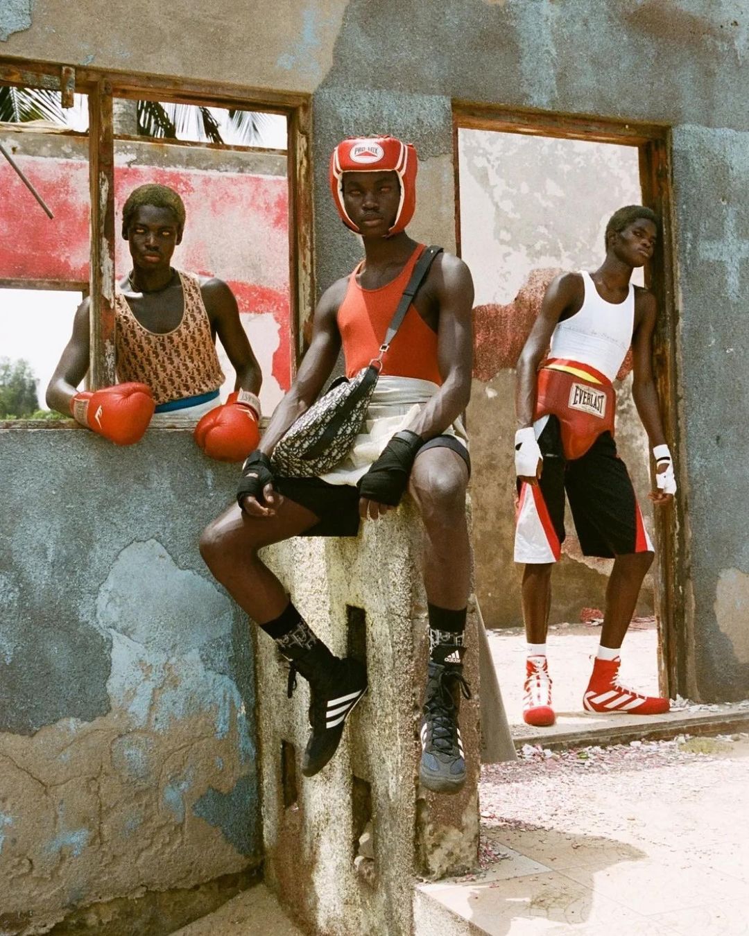Three male boxers pose inside an old stone structure with window openings, wearing boxing gear, shorts, and athletic shoes in an outdoor shot.