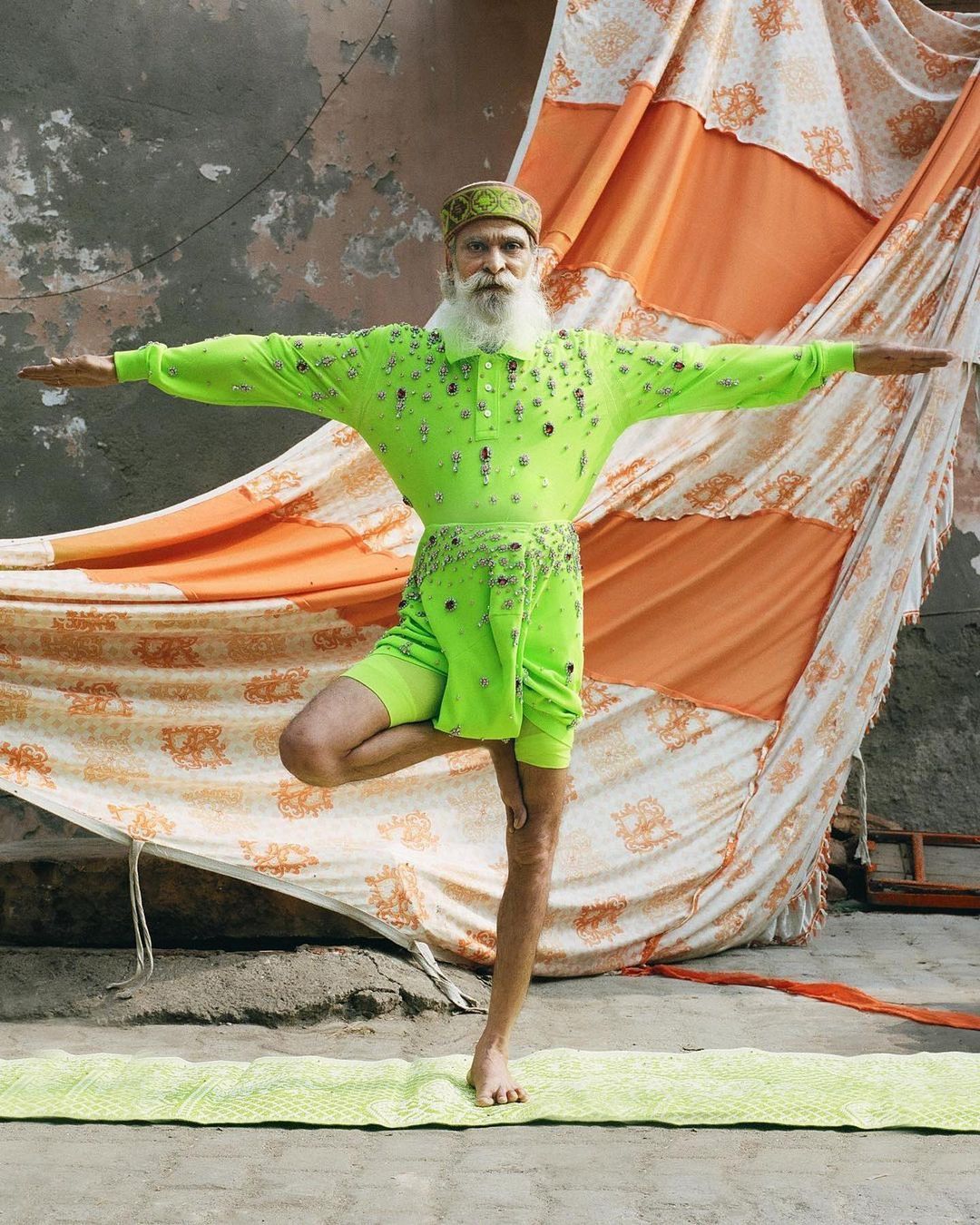 Elderly male yogi in bright green outfit balances in tree pose on a mat outdoors; orange and white fabric backdrop.
