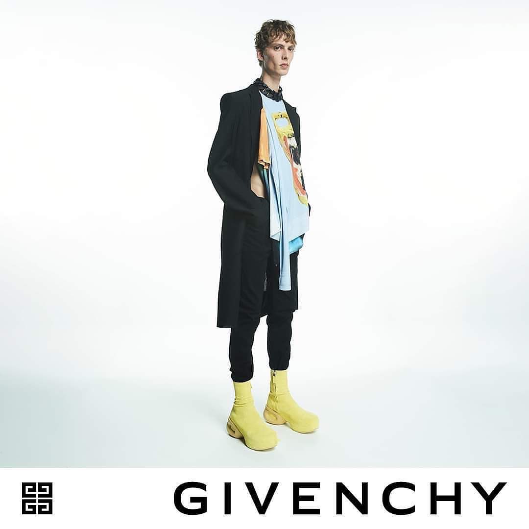 Male fashion model wearing black coat, graphic tee, black pants, and yellow boots stands in a brightly lit photo studio for Givenchy.