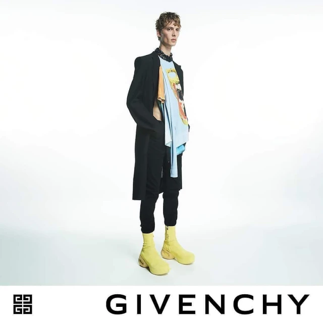 Male fashion model wearing black coat, graphic tee, black pants, and yellow boots stands in a brightly lit photo studio for Givenchy.