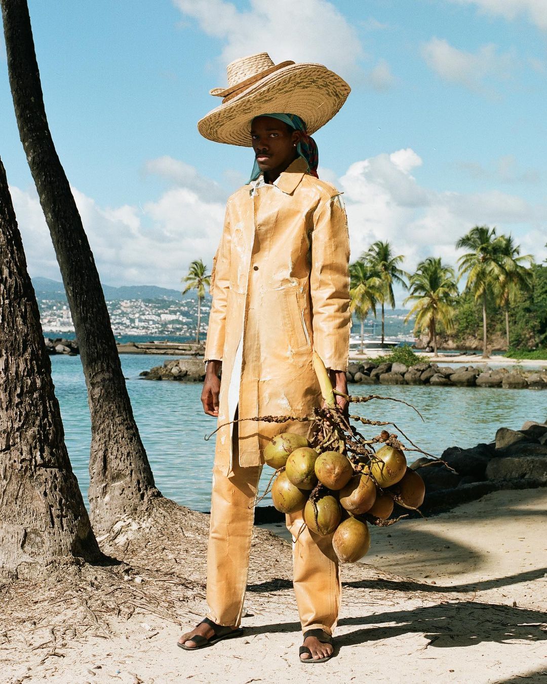 Young man in a tan coat and hat holding coconuts on a beach with palm trees and turquoise water under a bright blue sky.