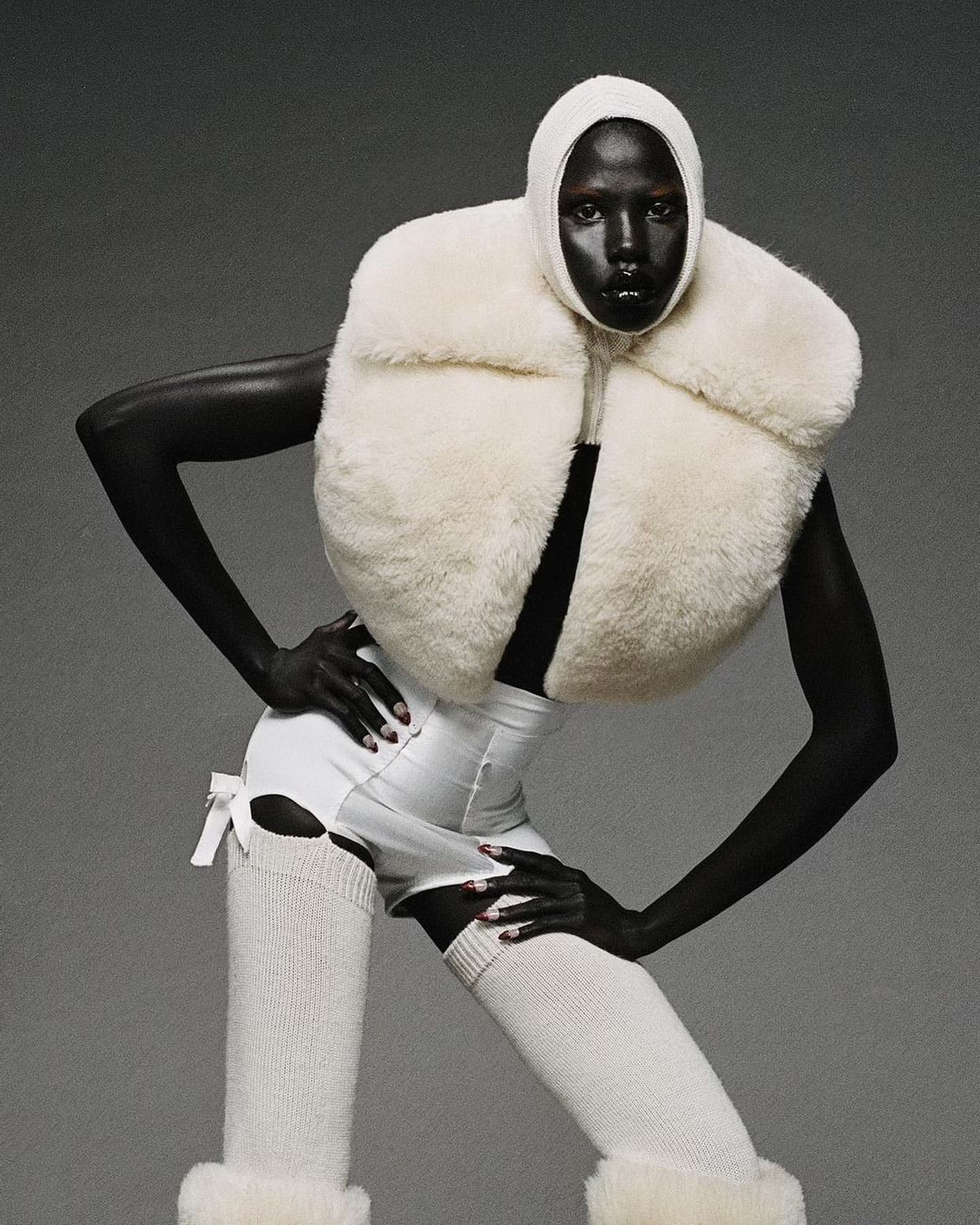 A woman with dark skin and a shaved head poses in an off-white fur vest, white high-waisted shorts, and thigh-high socks against gray.