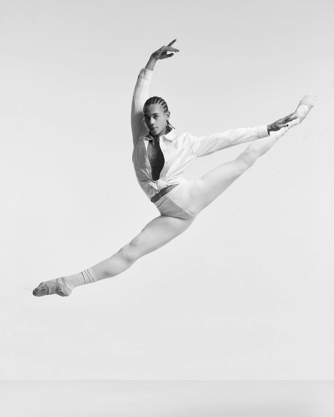 Black male ballet dancer mid-air in a split leap wearing white shirt and leggings against a light background in a dance studio.