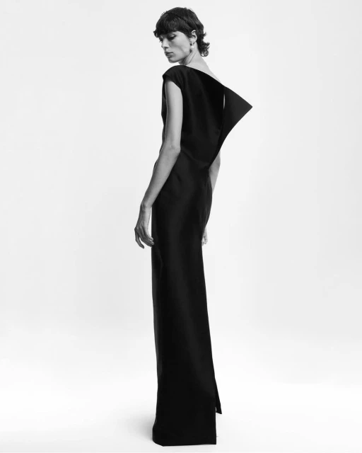 Black and white portrait of a model wearing a long, dark dress with a unique angular design, posing in a bright photo studio setting.
