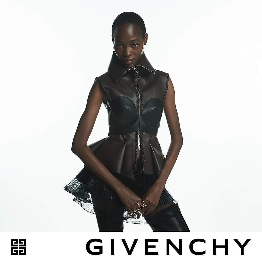 Fashion shoot of an adult African woman wearing a brown leather peplum top against a white background, jewelry on hands, and a Givenchy logo.
