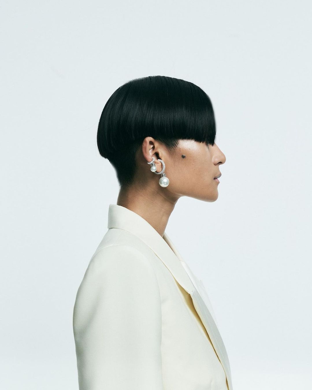 Profile view of a woman with a black bowl cut hairstyle wearing a white blazer and pearl drop earrings against a white background.
