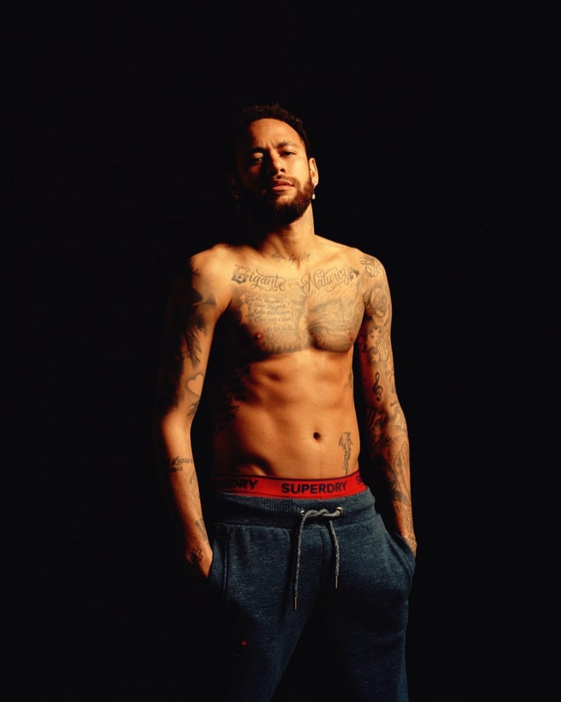 Portrait of shirtless man with tattoos, wearing sweatpants and a serious expression, set against a solid dark background.