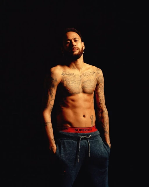 Portrait of shirtless man with tattoos, wearing sweatpants and a serious expression, set against a solid dark background.