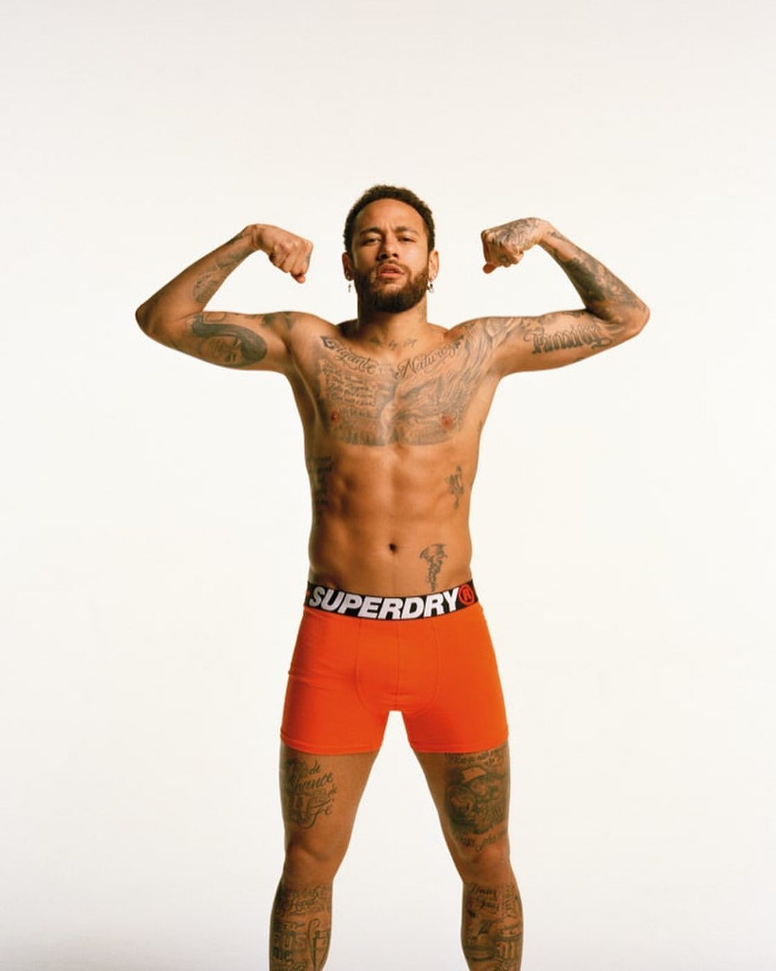 Studio shot of bare-chested man with arm tattoos flexing biceps, wearing orange Superdry underwear on a white backdrop.