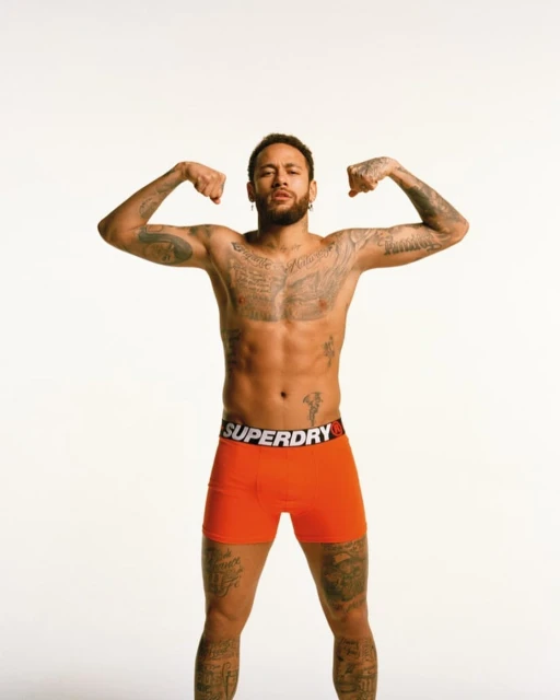 Studio shot of bare-chested man with arm tattoos flexing biceps, wearing orange Superdry underwear on a white backdrop.