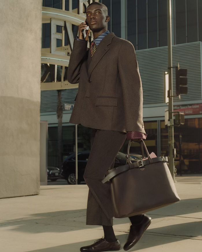 An African-American man in a brown suit walks through the city while talking on his cell phone, carrying a large brown tote bag.