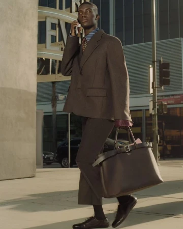 An African-American man in a brown suit walks through the city while talking on his cell phone, carrying a large brown tote bag.
