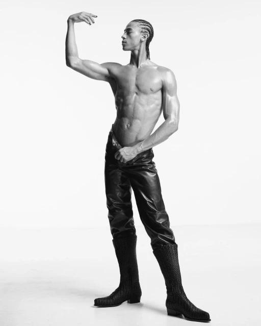 Black and white portrait of a muscular adult male, posing shirtless in leather pants and boots in a bright photo studio environment.