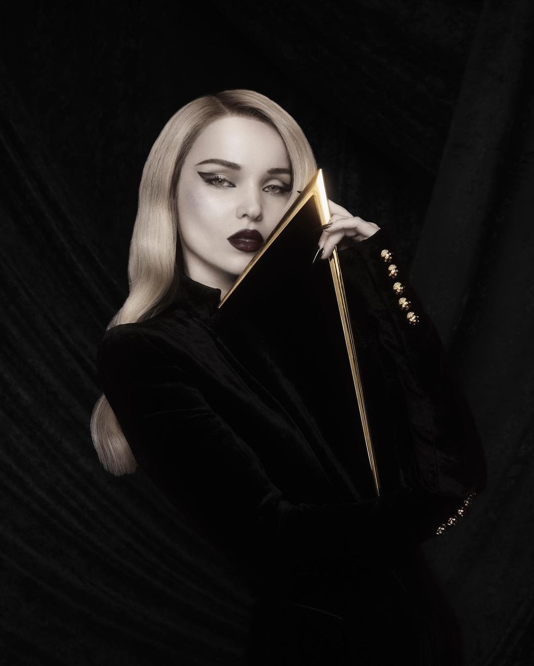 Glamorous woman with platinum blonde hair poses in black velvet jacket with gold accents, holding a black and gold clutch in a dark studio.