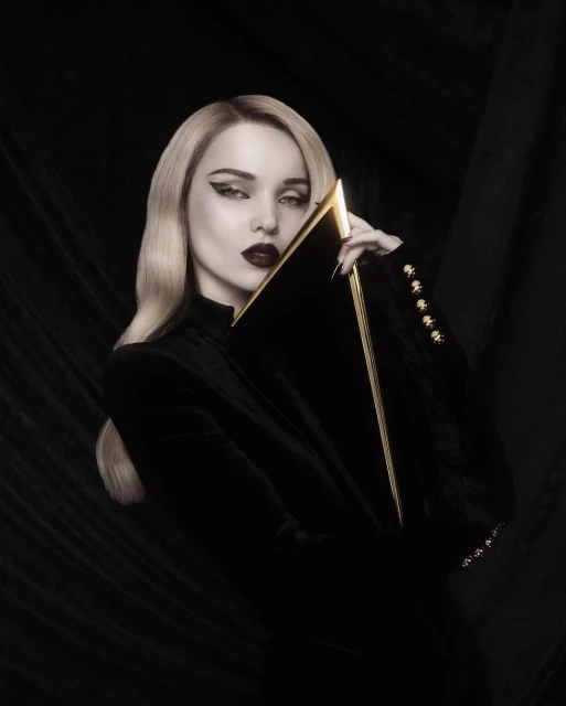 Glamorous woman with platinum blonde hair poses in black velvet jacket with gold accents, holding a black and gold clutch in a dark studio.