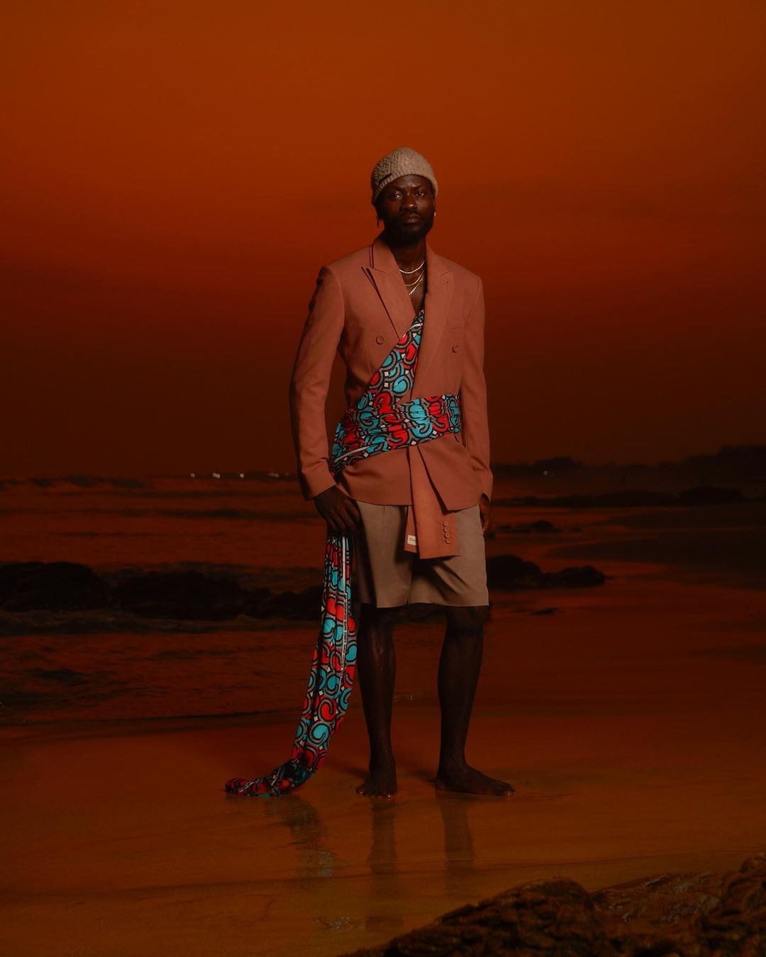 Man wearing a pink blazer, shorts, beanie, and patterned sash standing on a sandy beach at sunset.